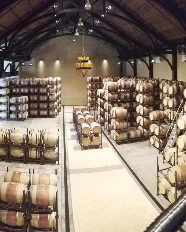 A large barrel room with oak barrels stacked in rows beneath an arched timber ceiling and a chandelier