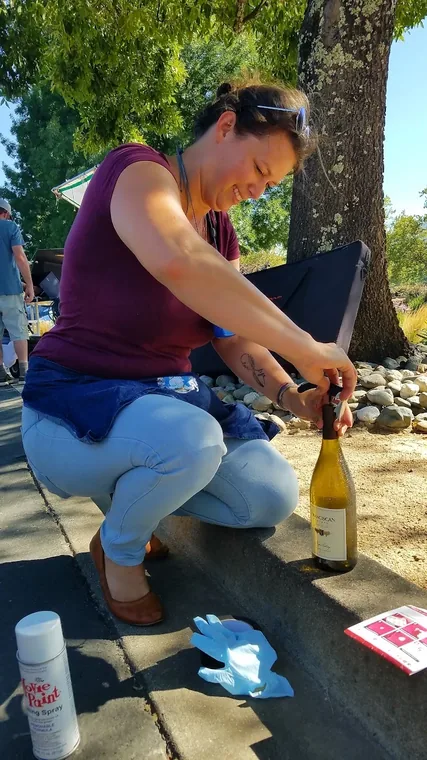 A stylist crouches on a path preparing a bottle of wine beside gloves and a spray can