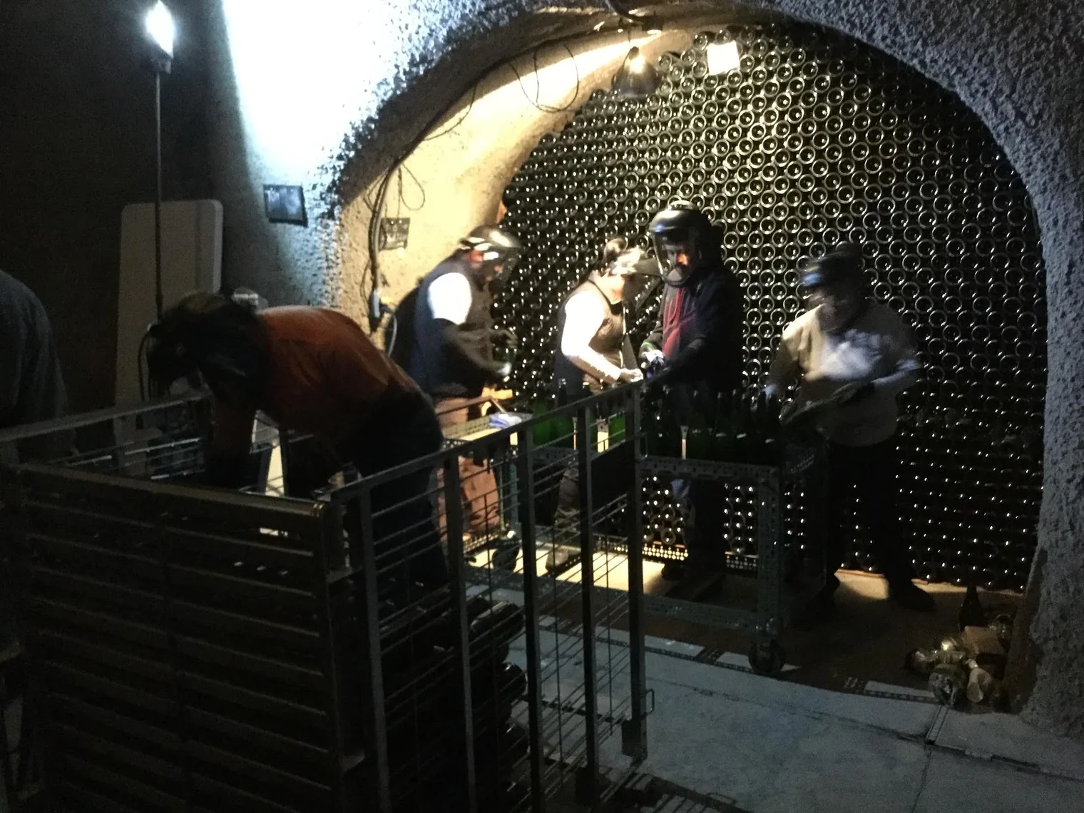 Winery workers in protective masks handling bottles at a rack inside a stone wine cave