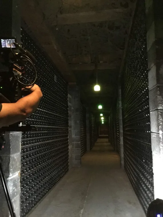 A camera operator films down a narrow cellar corridor lined floor to ceiling with stacked bottles