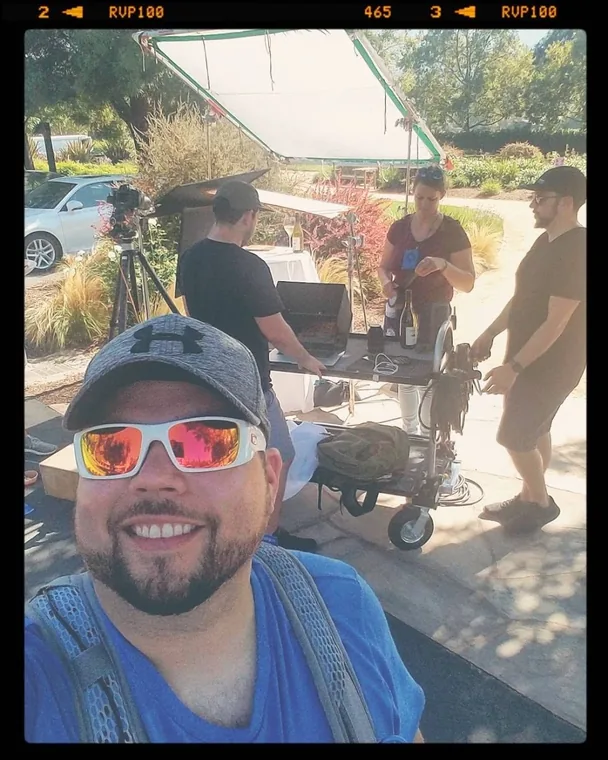 A crew member in sunglasses in the foreground while colleagues work at an outdoor food-styling setup behind
