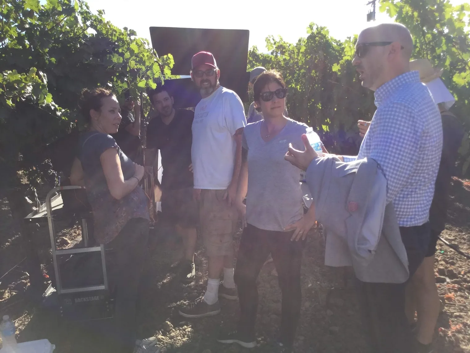 Crew members talking between takes in a sunlit vineyard with a reflector behind them