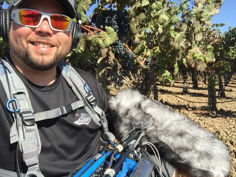 A sound recordist in headphones and sunglasses stands beside a vine row with a boom microphone and a mixer bag