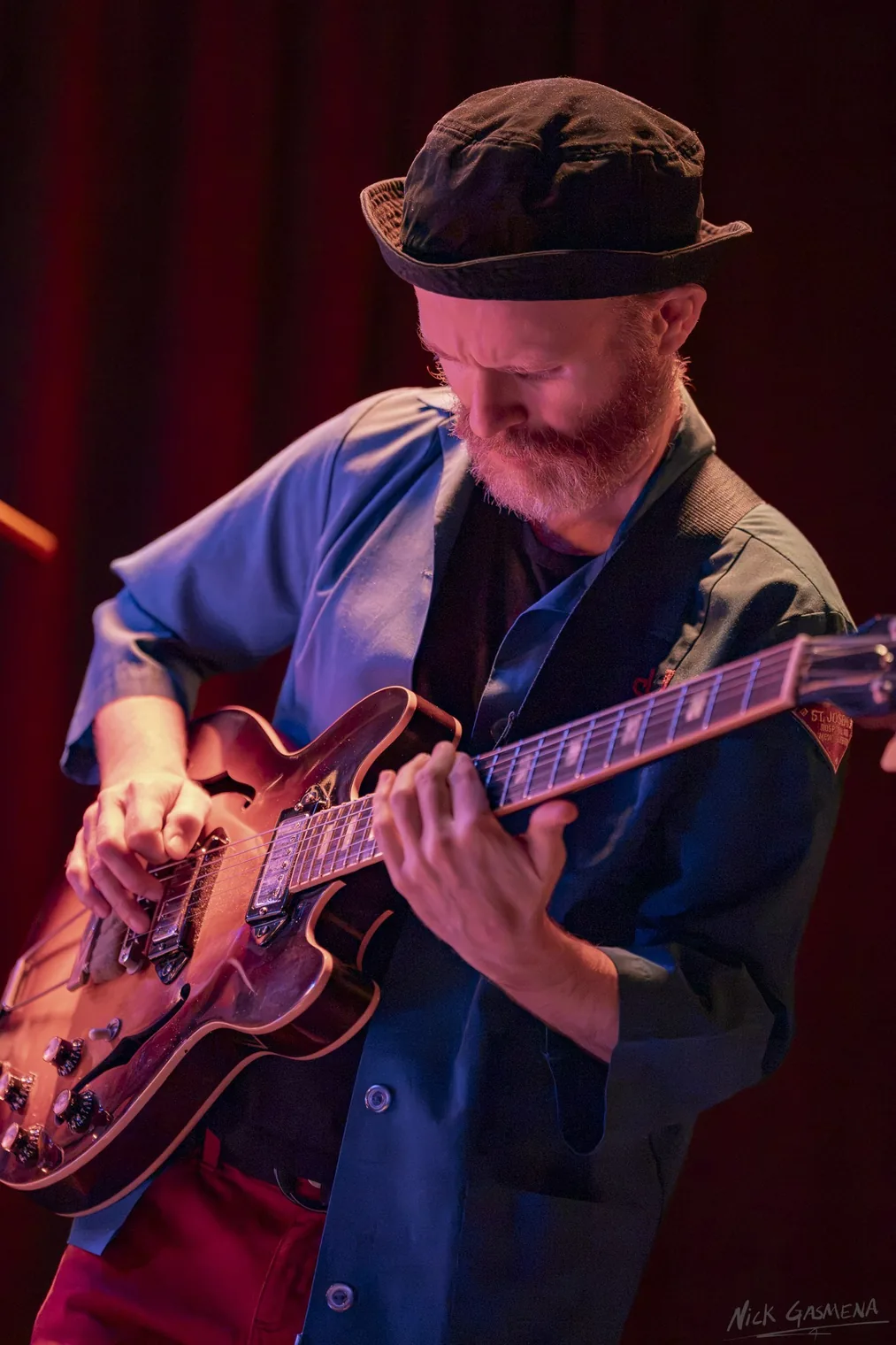 Ilan Smilan plays a hollow-body electric guitar, wearing a wide-brim hat, under warm stage light.