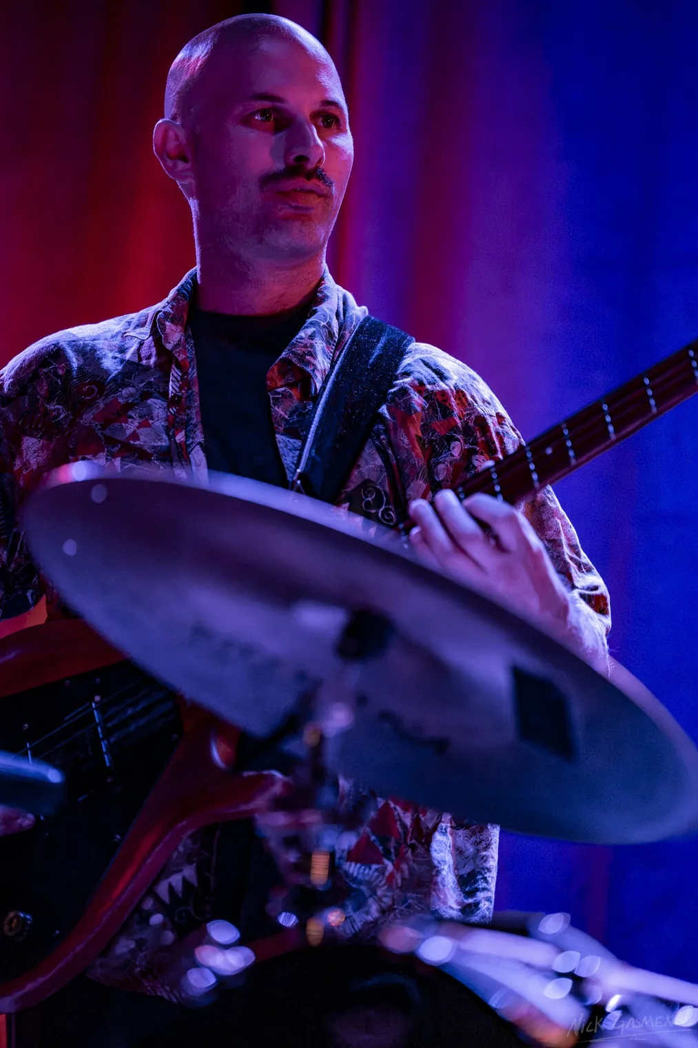 Amir Sadot plays bass guitar behind a cymbal, lit red and blue.