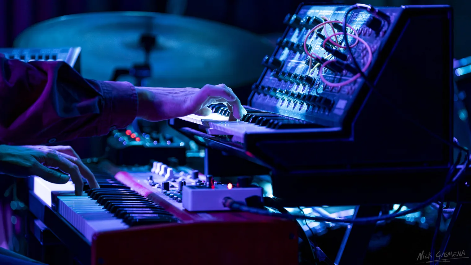 Hands play a patched analog synthesizer and keyboard under blue light.