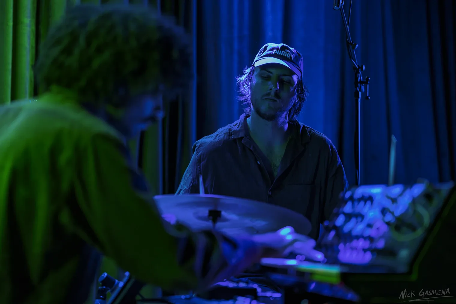 Itamar Weinstein plays drums under deep blue light, another musician blurred in the foreground.
