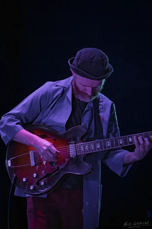 Ilan Smilan plays a hollow-body electric guitar under violet light, looking down at the strings.