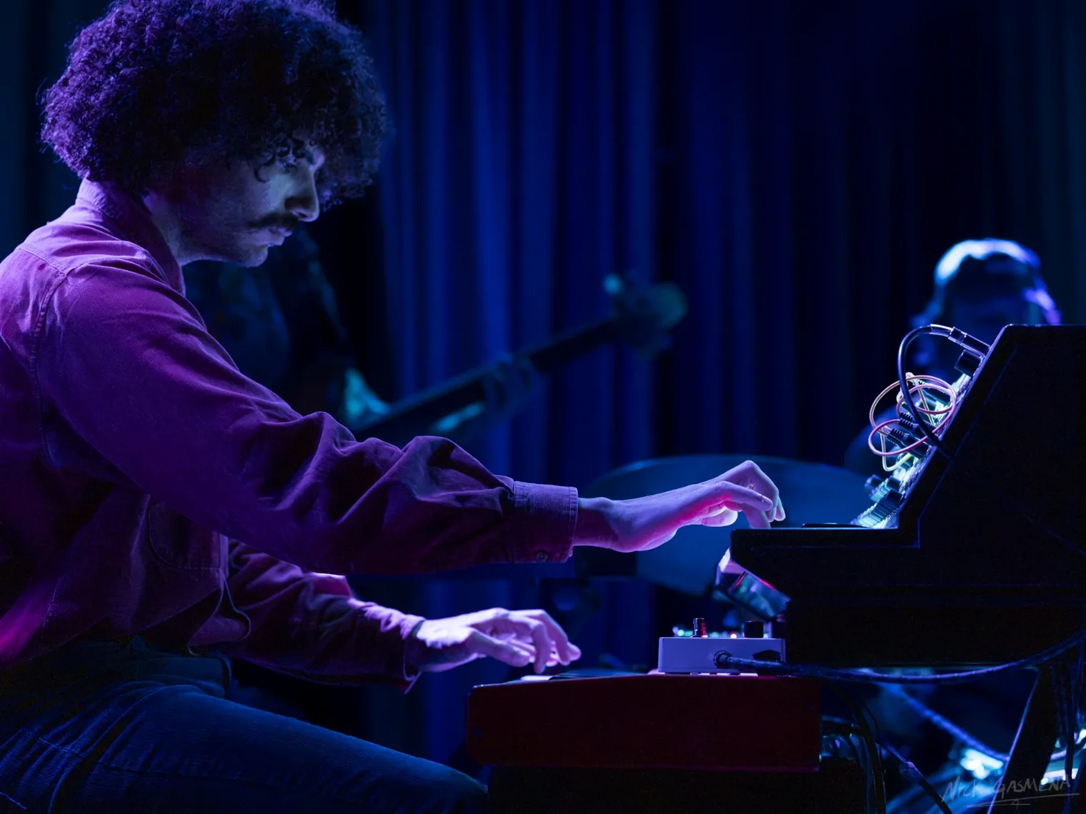 Eitan Drabkin plays two stacked keyboards, seen from the side, under blue and violet light.