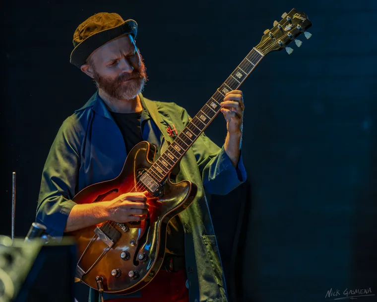 A guitarist in a cap and olive jacket plays a sunburst Gibson semi-hollow guitar against a dark blue wall