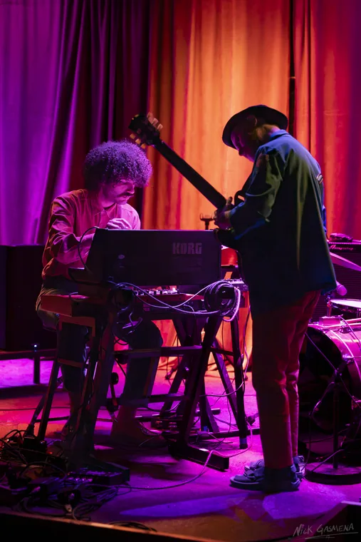 Eitan Drabkin seated at a KORG keyboard while Ilan Smilan leans over him with a guitar, framed against an orange and magenta curtain