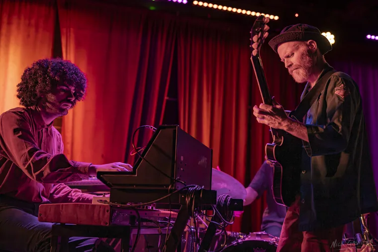 Eitan Drabkin plays keys at left while Ilan Smilan stands at right holding his guitar neck upright, both under deep red stage light