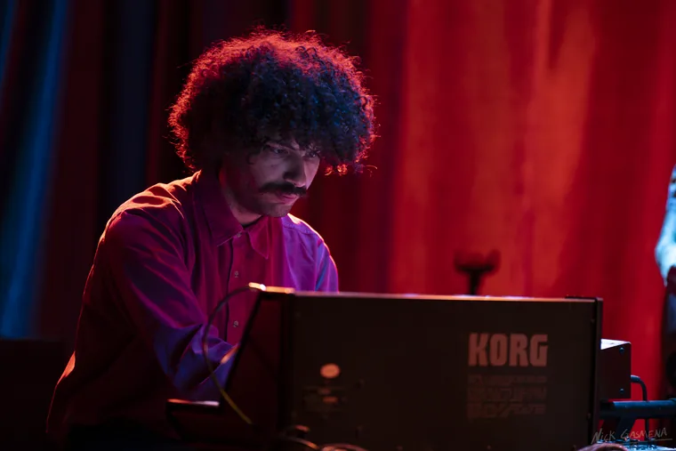 A keyboard player with dark curly hair and a moustache leans over a Korg synthesizer, lit deep red against a red stage curtain