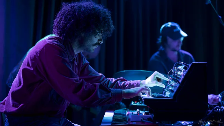 A keyboard player in profile under blue light with both hands on a synthesizer, the drummer in a cap visible behind him