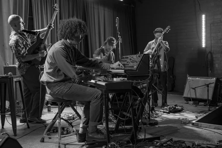 Black and white photograph of all four members of Sababa 5 playing on stage, bass at left, keys centre, drums and guitar at right