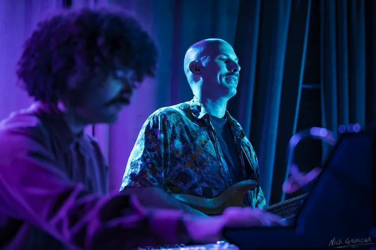 Amir Sadot sings and plays bass in a patterned shirt under blue light, with Eitan Drabkin blurred in the foreground