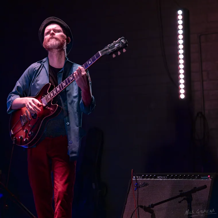 Ilan Smilan stands with a cherry semi-hollow guitar in a hat and red trousers, looking up, beside a vertical strip of stage lights